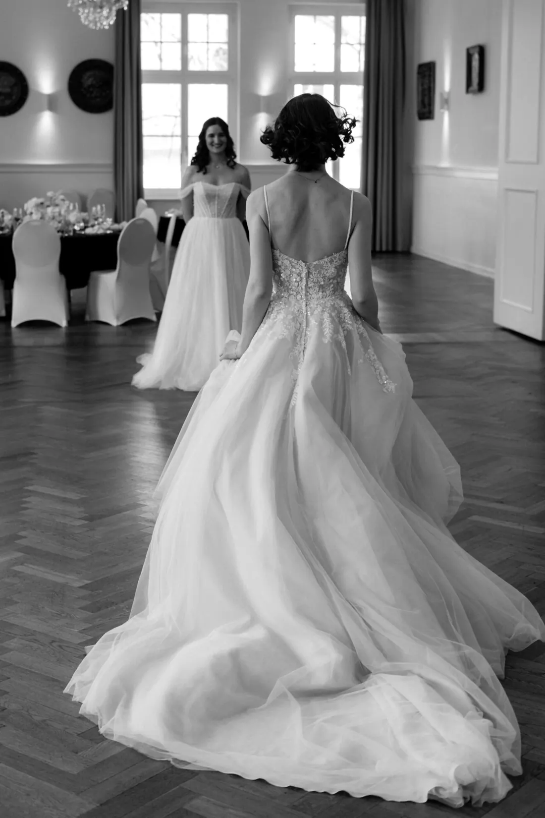 Braut im Tüllkleid geht durch einen festlich gedeckten Saal, im Hintergrund eine zweite Braut